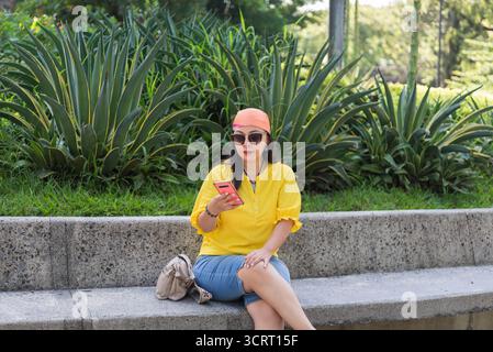 Une femme en chemise jaune et foulard orange est assise sur un banc de parc, utilisant son téléphone. Banque D'Images
