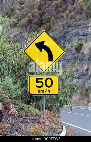 Limite de vitesse jaune et tourner à gauche devant les panneaux routiers parmi les arbres et une falaise Banque D'Images