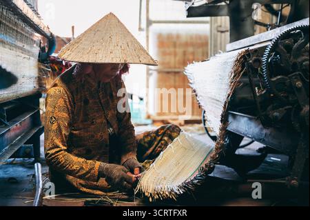 Femme vietnamienne tisserand en chapeau de paille travaille aux machines à tisser à métier dans l'atelier à l'usine pour la production de nattes tissées de roseau au Vietnam. An Cu, Tuy an, province de Phu Yen, Vietnam - 14 avril 2025 Banque D'Images