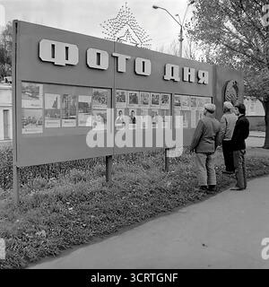 Une authentique photographie d'archives en noir et blanc des années 1980 montre un groupe d'hommes à Sloviansk, en RSS d'Ukraine, lisant un panneau d'information publique. Le grand stand, intitulé « photo du jour » (« Фото дня » en cyrillique), était une forme courante de médias de rue soviétiques et d'« agitation visuelle ». Il affiche des photographies officielles de nouvelles, des portraits de travailleurs exemplaires et des informations politiques pour les citoyens. Cette scène de rue franche est une illustration puissante de la façon dont les nouvelles et la propagande contrôlées par l'État étaient consommées dans la vie quotidienne en URSS Banque D'Images