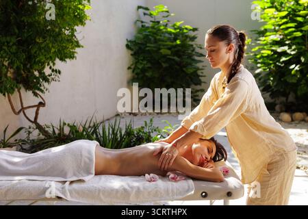 Le thérapeute effectue un massage relaxant du dos en plein air dans le jardin du spa, la lumière naturelle et les orchidées transmettent bien-être, calme et hospitalité haut de gamme. Banque D'Images