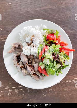 Un repas nutritif avec des cœurs de poulet mijotés et gésiers, du riz et de la salade fraîche est servi dans l'assiette. Porte-plats sur une table en bois rustique Banque D'Images