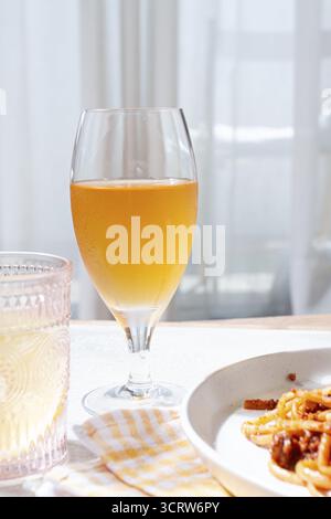 Un verre rempli d'une boisson ambre légère se trouve à côté d'une assiette de pâtes. La lumière vive du soleil traverse des rideaux transparents, créant un guichet automatique chaleureux et accueillant Banque D'Images