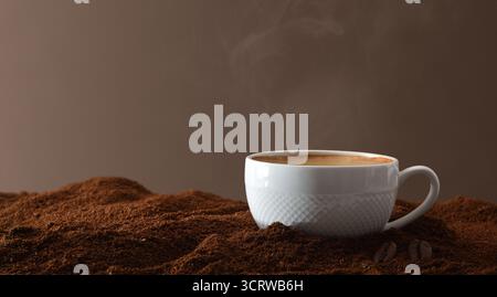 Tasse en porcelaine blanche avec du café chaud fraîchement infusé sur un tas de café moulu et des grains autour avec un fond isolé brun. Vue de face. Banque D'Images