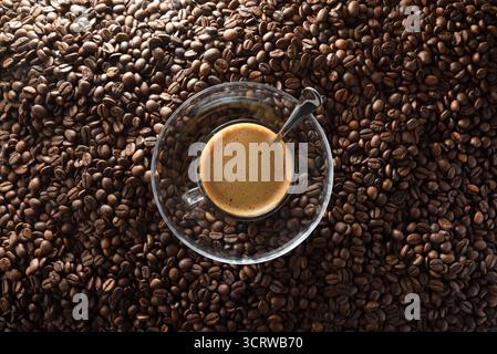Tasse de café mousseux sur une assiette en verre avec une cuillère en métal au centre sur le dessus d'un tas de grains de café torréfiés. Vue de dessus. Banque D'Images