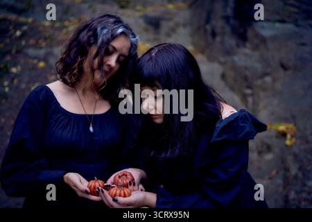 Deux femmes en robes longues noires près des rochers noirs sur Halloween avec des bougies en forme de citrouilles, effectuant un rituel de sorcière, sorcière moderne Banque D'Images