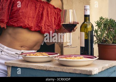 Une femme méconnaissable dans un sommet rouge tient un verre de vin rouge, profitant d'un simple déjeuner rustique de spaghettis sur un balcon extérieur en Italie. Banque D'Images
