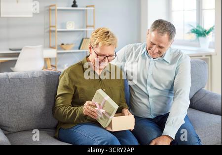 Heureux couple senior assis sur le canapé à la maison avec boîte cadeau cadeau célébrant les vacances. Banque D'Images