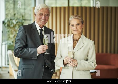 Homme caucasien senior et femme caucasienne senior debout ensemble tenant des verres de champagne, souriant à la caméra dans un cadre intérieur moderne, célébrant une occasion spéciale Banque D'Images