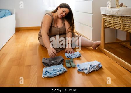 Femme enceinte triant les vêtements de bébé dans la pépinière sur plancher en bois. Style de vie. Grossesse. Banque D'Images