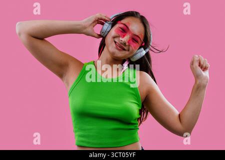 Jeune femme souriante et dansante, écoutant de la musique sur ses écouteurs, portant des lunettes de soleil en forme de cœur et un débardeur vert sur un bac rose éclatant Banque D'Images