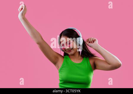 Jeune femme souriante et dansante, écoutant de la musique sur ses écouteurs, portant des lunettes de soleil en forme de cœur et un débardeur vert sur un bac rose éclatant Banque D'Images