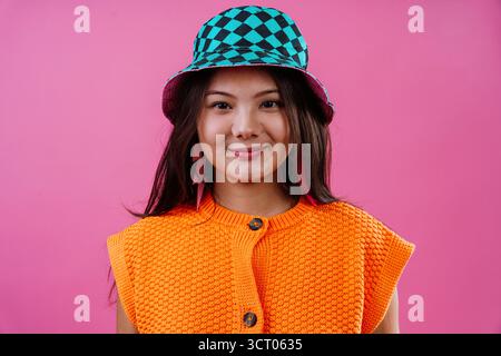 Jeune femme souriant à la caméra, portant un pull en tricot orange et un chapeau seau à carreaux bleu sarcelle et noir éclatant, debout contre un BA rose vif Banque D'Images
