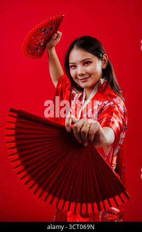Jeune femme asiatique portant un kimono rouge traditionnel avec des motifs floraux, tenant deux ventilateurs de main rouges, et souriant à la caméra contre un BA rouge vif Banque D'Images