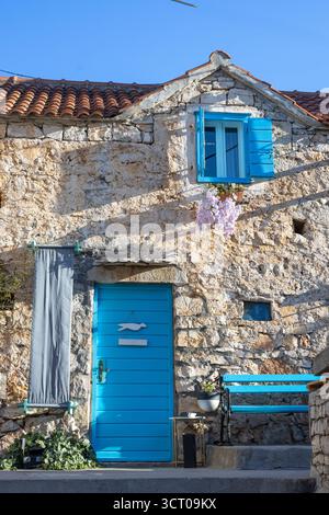 Une maison en pierre altérée avec des portes et des volets bleus vifs se trouve sur une rue méditerranéenne ensoleillée. Ivy grimpe le mur, un banc bleu ajoute du charme, et Banque D'Images