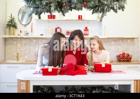Famille ouvrant des cadeaux de Noël dans la cuisine festive. Une mère souriante et ses deux filles se réunissent autour d’une table de cuisine décorée pour Noël Banque D'Images