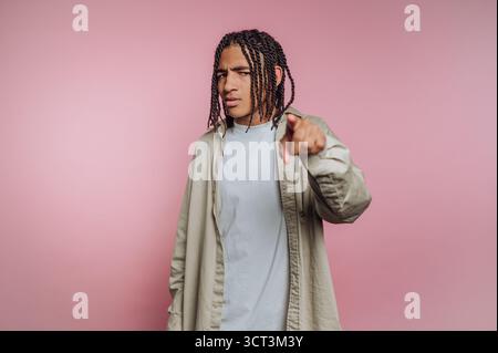 Un jeune homme se tient confiant avec un regard sérieux, pointant directement vers le spectateur sur un fond rose Uni. Il porte une tenue décontractée qui ajoute Banque D'Images