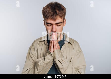 Un jeune homme se tient dans un espace intérieur bien éclairé, les yeux fermés et les mains pressées ensemble dans un geste de contemplation ou de prière. Il semble concentré et Banque D'Images