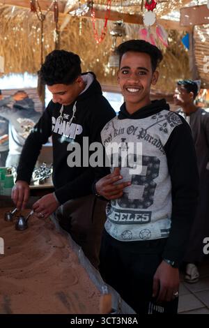 Sinaï Sud, Egypte - 5 février 2024 : Un adolescent bédouin arabe joyeux et son ami font du café sur le sable pour les touristes. Un village dans le Sinaï mou Banque D'Images
