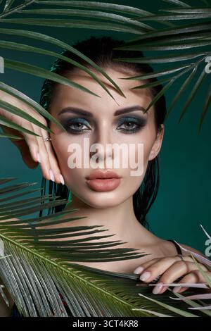 femme au maquillage saisissant pose devant des feuilles vertes vibrantes. Ses doigts touchent doucement son visage tandis qu'elle maintient un regard captivant. backgroun Banque D'Images