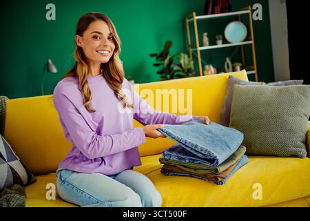 Attrayante jeune femme pliant des vêtements à la maison sur une journée joyeuse assise sur un canapé vibrant dans un appartement confortable Banque D'Images