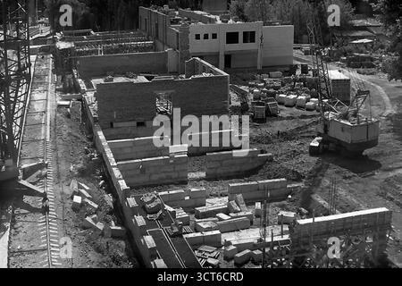 Une vue en grand angle d'un grand chantier de construction à Sloviansk, en RSS d'Ukraine, au cours des années 1980 Cette photographie d'archives capture le développement urbain à la fin de l'ère soviétique, montrant la phase initiale de construction de ce qui est probablement un nouveau 'Univer-sam' (supermarché). La fondation est posée avec de grands blocs de béton et les premiers murs de briques se dressent. Les grues à tour et mobiles dominent la scène, entourées de matériaux de construction, illustrant les méthodes industrielles de l'époque. Cette image de création active symbolise la croissance paisible et la modernisation de la ville Banque D'Images