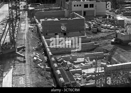 Une vue en grand angle d'un grand chantier de construction à Sloviansk, en RSS d'Ukraine, au cours des années 1980 Cette photographie d'archives capture le développement urbain à la fin de l'ère soviétique, montrant la phase initiale de construction de ce qui est probablement un nouveau 'Univer-sam' (supermarché). La fondation est posée avec de grands blocs de béton et les premiers murs de briques se dressent. Les grues à tour et mobiles dominent la scène, entourées de matériaux de construction, illustrant les méthodes industrielles de l'époque. Cette image de création active symbolise la croissance paisible et la modernisation de la ville Banque D'Images