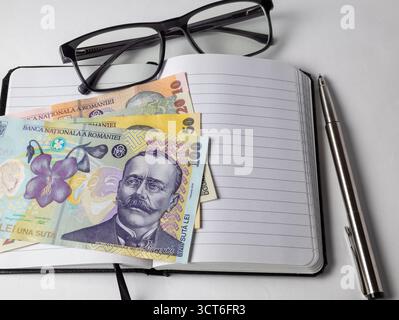 gros plan de billets roumains en lei (10, 50, 100) sur un bloc-notes ouvert avec un stylo élégant et des lunettes de lecture noires, symbolisant la budgétisation et la volonté financière Banque D'Images