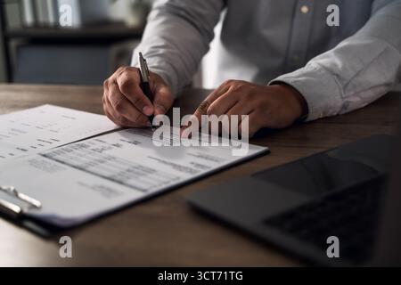 Concept de signature de contrat pour l'approbation financière : gros plan des mains examinant et signant les documents sur une planchette à pince au bureau, avec un ordinateur portable à proximité, SY Banque D'Images