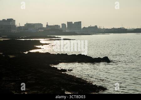 La lumière de fin de journée scintille le long de la ligne de flottaison à Sehwa tandis que les bas promontoires de lave noire entrent dans la baie et que la ligne d'horizon du port se trouve dans une silhouette douce. Banque D'Images