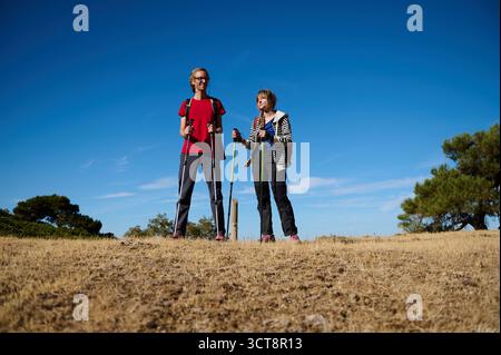 Deux amis marchent côte à côte avec des bâtons de trekking dans un paysage sec et ouvert sous un ciel bleu vif, véhiculant aventure en plein air, fitness et compagnon Banque D'Images