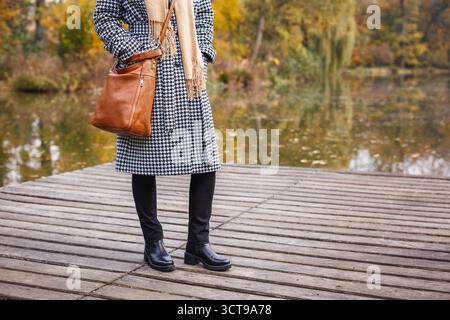Femme à la mode portant un manteau pied-de-poule et un pantalon noir debout près de l'étang dans le parc d'automne. Mode de vie de la saison d'automne Banque D'Images