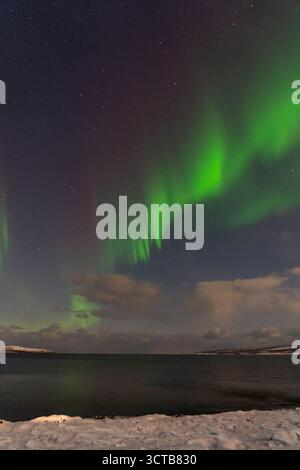 Mourmansk, Russie - 13 décembre 2024 : rubans verts vifs et intenses de l'aurore boréale s'arquant haut dans le ciel nocturne au-dessus d'un corps sombre et calme de wa Banque D'Images