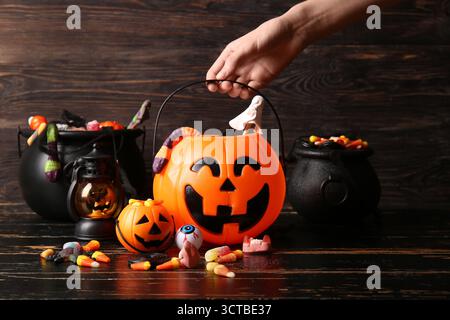 Main féminine avec des citrouilles décoratives et des chaudrons de différents bonbons sucrés sur une table en bois noir. Célébration d'Halloween Banque D'Images