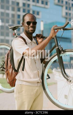 Jeune homme noir adulte souriant tout en transportant un vélo à l'extérieur dans la zone urbaine Banque D'Images
