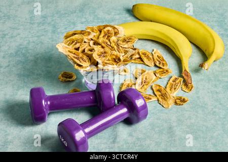 Concept de mode de vie sain : haltères, bananes fraîches et chips de bananes séchées pour l'énergie et la motivation de l'exercice sur une surface texturée Banque D'Images