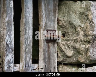 Loquet en métal oxydé sur vieux bois et pierre - gros plan d'un boulon à glissière fortement rouillé contre des planches de bois altérées et une texture de mur de pierre rugueuse. Banque D'Images