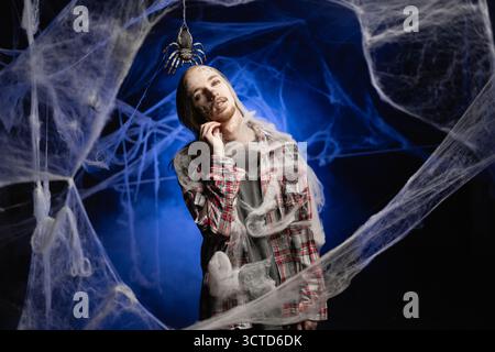 Mec mort avec horrible maquillage de zombie effrayant Halloween sur le visage essayant d'effrayer debout couvert de toiles d'araignées sur fond bleu. copier l'espace Banque D'Images
