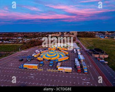 Vue aérienne de la configuration vibrante du cirque dans le parking éclairé au coucher du soleil tente bleu jaune, les remorques et les camions contrastent avec le ciel rose et la toile de fond boisée, ble Banque D'Images