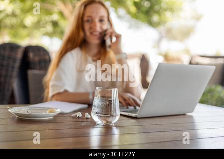 Une jeune femme caucasienne aux longs cheveux blonds travaille à distance dans un café. Elle est au téléphone, avec un ordinateur portable et un verre d'eau sur la table. Banque D'Images