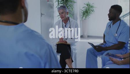 Médecin senior guidant la discussion d'équipe dans le salon de l'hôpital, avec bloc-notes et tablettes Banque D'Images