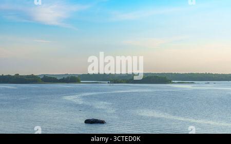 Lac calme avec des rochers dispersés près de la rive, entouré d'arbres verts et forêt lointaine sous un ciel bleu clair sur une journée d'été tranquille Banque D'Images