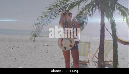 Tenant chapeau de paille et touchant les cheveux, femme portant des lunettes de soleil posant sur la plage de sable par hamac Banque D'Images