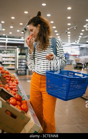 Femme en chemise rayée inspecte une tomate rouge mûre dans l'allée des produits du supermarché, sentant la fraîcheur tout en sélectionnant des légumes sains et vibrants pendant les courses Banque D'Images