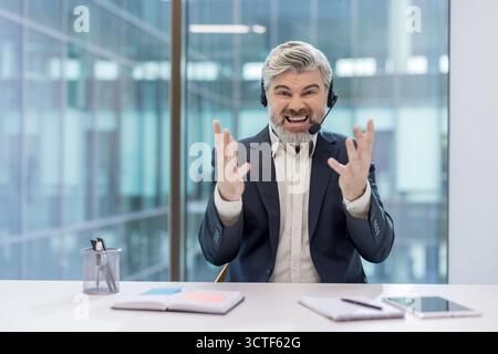 Agent de centre d'appels frustré criant dans le micro-casque au bureau, affichant une colère et un épuisement visibles lorsqu'il traite un client difficile, illustrant le stress et la tension sur le lieu de travail Banque D'Images