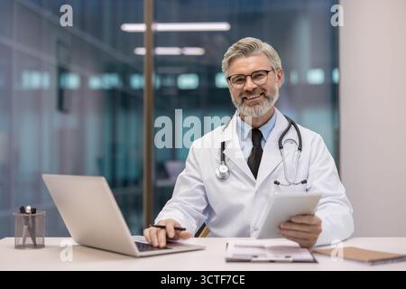 Homme médecin mûr souriant, portant un stéthoscope et un manteau blanc, travaillant dans son bureau avec un ordinateur portable et une tablette numérique, montrant l'innovation et les soins de santé modernes Banque D'Images