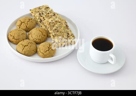 Une assiette blanche avec des biscuits à l'avoine et des craquelins de graines servis à côté d'une tasse de café noir sur un fond blanc. Concept minimaliste de petit-déjeuner ou de snack Banque D'Images