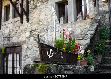 Yvoire, France - 25 septembre 2025 : un panier décoratif en osier en forme de bateau, rempli de fleurs vibrantes, placé sur un mur de pierre devant un Banque D'Images