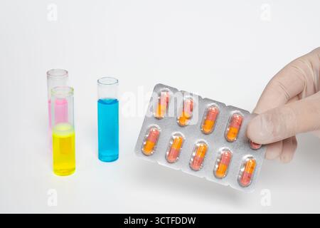 Main gantée tenant un blister de capsules orange et jaune avec des tubes à essai colorés sur fond blanc isolé. La recherche médicale avec les pilules Banque D'Images