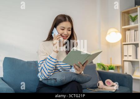 Une jeune femme assise confortablement sur un canapé, souriante tout en utilisant un smartphone avec un ordinateur portable sur ses genoux, montrant une scène de multitâche entre digi Banque D'Images